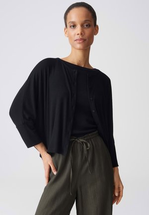 Frau trägt eine locker sitzende schwarze Strickjacke mit Knöpfen über einem schwarzen Oberteil und olivgrünen Kordelhosen, sie steht vor einem einfachen hellen Hintergrund.