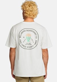 Camiseta de algodón blanca con un gráfico redondo en la parte posterior que muestra texto verde y naranja y un motivo de planta. Mangas cortas, corte relajado.