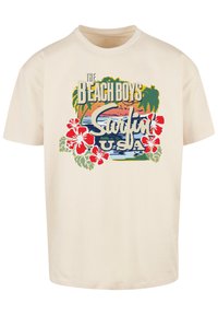 Cream-colored T-shirt featuring a graphic design with tropical elements, including hibiscus flowers, palm trees, and sunset colors.