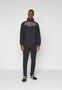 Fekete sportdzseki, katonai mintás nyakkal, fekete, slim-fit nadrágon ül. A modellt fekete és arany lábbeliben látjuk.