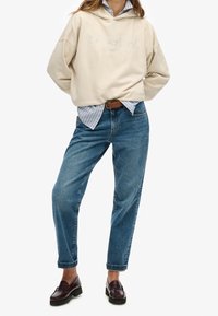 Sudadera beige con el logo en relieve de "SuperDry", superpuesta sobre una camisa a rayas azul y blanca, combinada con unos vaqueros holgados azules y mocasines oscuros.