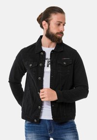 Veste en denim noir avec un design à boutons sur le devant, deux poches poitrine et des poignets réglables. Portée par-dessus un t-shirt graphique blanc.
