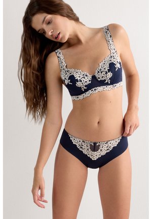Femme portant un ensemble de lingerie bleu marine avec des détails en dentelle florale blanche, posant la tête inclinée et la main sur la hanche sur un fond blanc uni.
