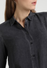 Donna che indossa una camicia grigia scura con bottoni e colletto, mostrata dalle spalle al mento, con la testa leggermente girata di lato.