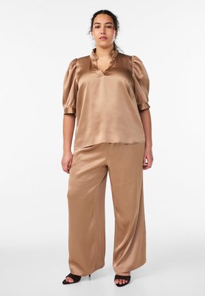 Kvinde iført en beige, silkeagtig bluse med pufærmer og matchende viddebukser, stående foran en ensfarvet hvid baggrund.