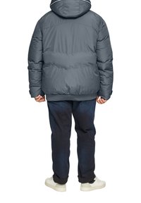 Donkergrijze pufferjack met een capuchon, voorzien van een gestikte textuur en elastische manchetten, gecombineerd met donkere jeans en witte sneakers.