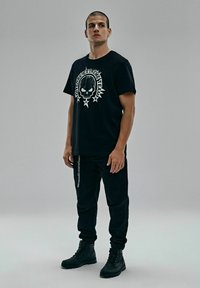 Czarna koszulka z nadrukiem czaszki i tekstem, w połączeniu z czarnymi spodniami i solidnymi czarnymi butami. Styl casualowy, streetwear.