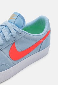 Hellblaue Wildledersneaker mit einem leuchtend orangefarbenen Nike-Swoosh, strukturierter Gummisohle und grünem Innenfutter. Die Schnürsenkel sind straff gebunden.