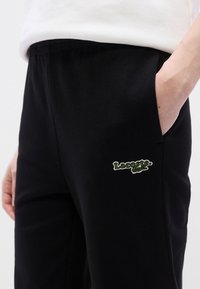 Μαύρο παντελόνι φόρμας με ελαστική μέση, πλαϊνές τσέπες και πράσινο κεντημένο λογότυπο Lacoste στο αριστερό μηρό. Απαλό βαμβακερό ύφασμα.