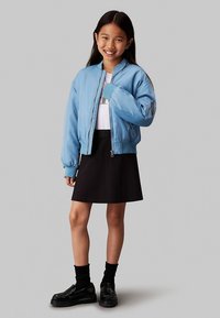 Svetlo modra bomber jakna z rebrastimi manšetami, nošena čez belo srajco in črno krilo. Model nosi črne čevlje in nogavice, stoji na nevtralnem ozadju.