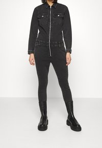 Combinaison en denim noir à manches longues, fermeture éclair, deux poches poitrine, jambes ajustées et taille élastique, associée à des bottines noires.