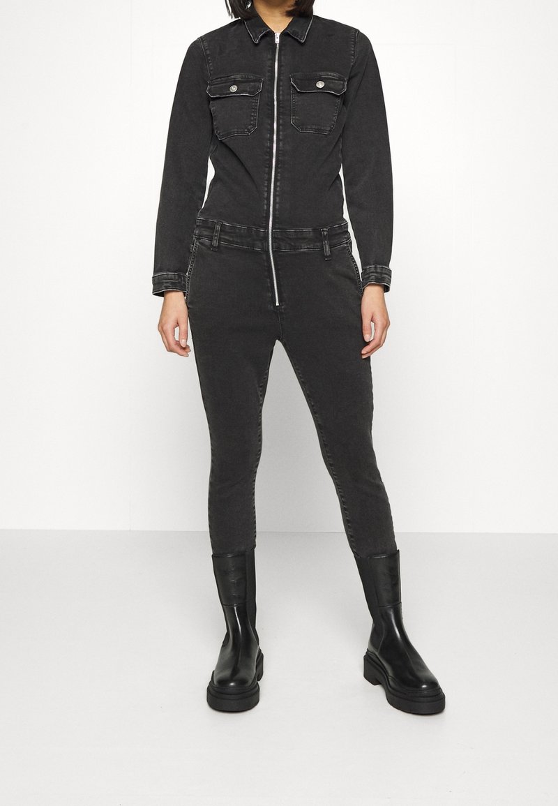 Combinaison en denim noir à manches longues, fermeture éclair, deux poches poitrine, jambes ajustées et taille élastique, associée à des bottines noires.