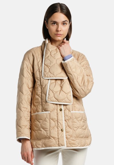 Giacca trapuntata beige con grande colletto, piping bianco e due tasche frontali. Il tessuto è liscio e con una finitura lucida.