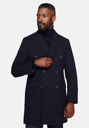 Man wearing a dark navy double-breasted coat and turtleneck, adjusting the coat with one hand, looking to the side against a plain background.