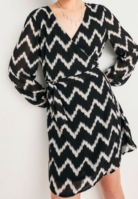 Svartvit chevron-inspirerad klänning med lång ärm, djup V-ringning och knytning i midjan. Lättviktsmaterial med en slät textur och svävande design.