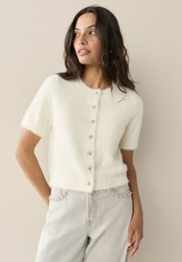 Cardigan bianco a maniche corte realizzato in materiale soffice, con scollo rotondo e chiusura frontale con bottoni decorativi a forma di gemma.