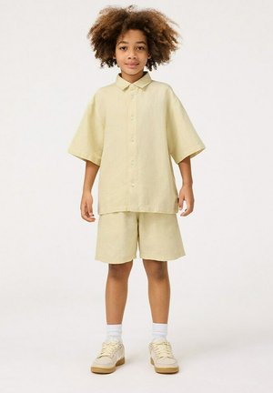 Camicia di lino gialla chiara con maniche corte e bottoni, abbinata a pantaloni corti coordinati. Indossata con calzini bianchi e sneakers color sabbia chiaro.