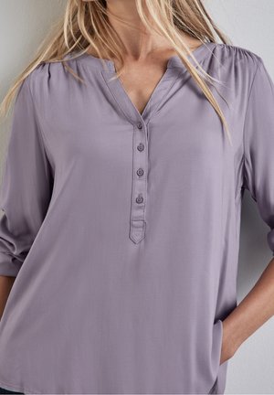 Kobieta nosząca luźną, jasnofioletową bluzkę z czteroprzyciskowym mankietem i rękawami do łokcia, ręka swobodnie w kieszeni.