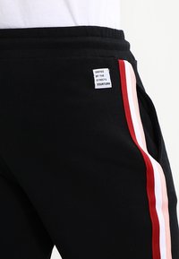 Zwarte sweatpants met verticale zijstrepen in rood, wit en roze en een klein wit etiket met de tekst "Unified by the streets Yourturn."