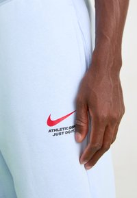 Gaiši zili sporta bikses, kas izgatavotas no mīksta auduma, ar sarkanu Nike logo un melnu tekstu "ATHLETIC DEPT. JUST DO IT." sānos.