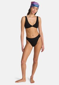 Vrouw staat op blote voeten, draagt een zwarte bikini en een paarse hoofddoek met patroon, tegen een witte achtergrond.