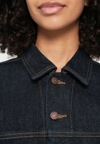 Kurtka dżinsowa z ciemnoniebieskiego materiału, z ostrym kołnierzem, brązowymi guzikami i kontrastującymi żółtymi przeszyciami. Zbliżenie na obszar kołnierza.