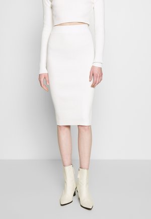 Kobieta ubrana w białą prążkowaną midi spódnicę ołówkową, długi crop top z długim rękawem oraz kremowe botki na obcasie, stojąca na jednolitym tle.