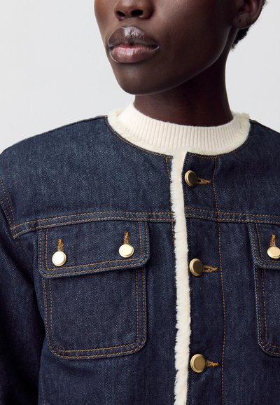 Giacca di jeans blu scuro con colletto e fodera a coste color crema. Presenta chiusure con bottoni dorati e due tasche anteriori a soffietto. Tessuto testurizzato visibile.