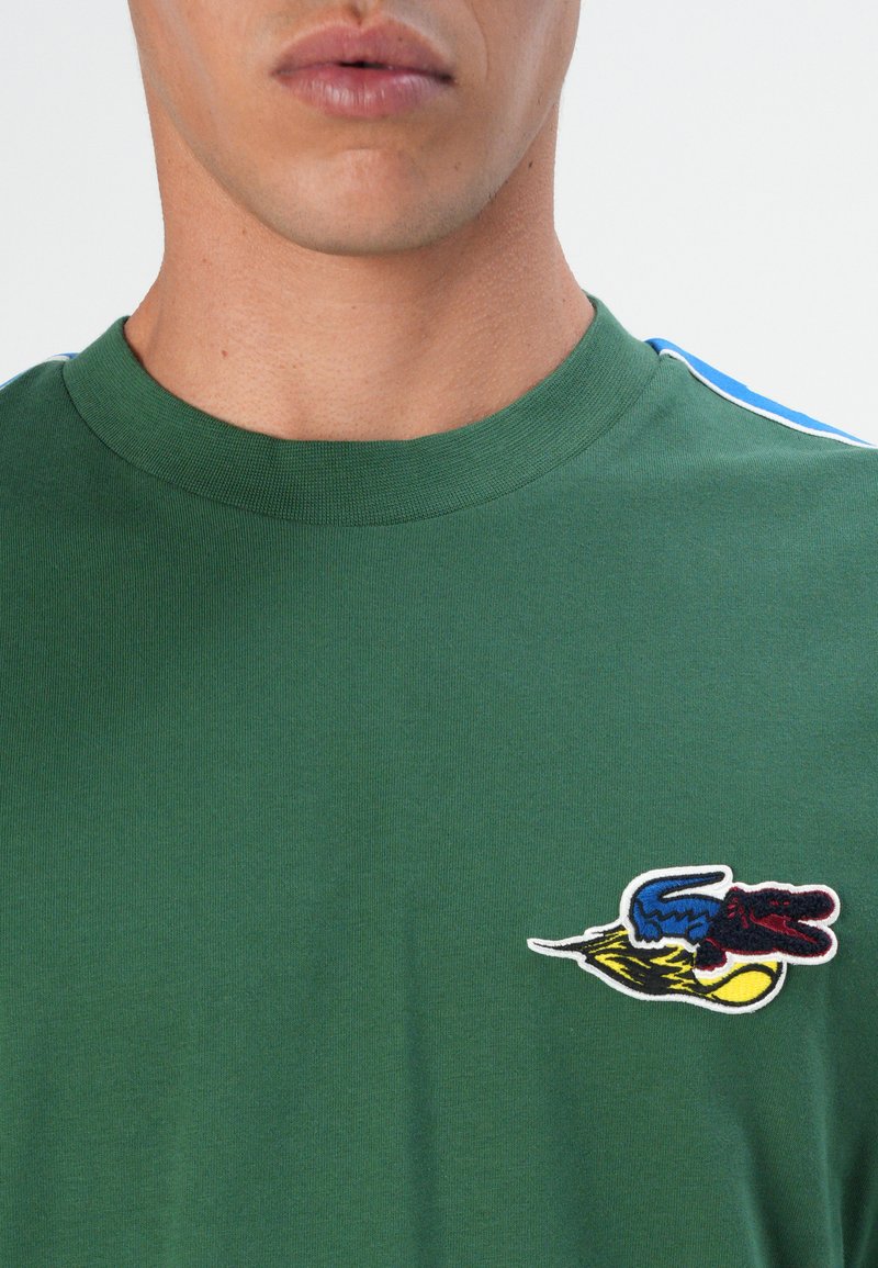 Zielona koszulka z krótkim rękawem, wykonana z bawełny, z wielokolorowym haftowanym logo na klatce piersiowej i niebieskimi akcentami na ramionach.