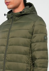 Olive green puffer jacket with a hood, featuring quilted sections, a front zipper, and a circular logo patch on the left sleeve.