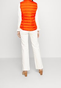 Vadderad, klar orange väst med vertikal quiltning, bärs över en vit långärmad topp, i kombination med krämfärgade vida byxor och träklackade sandaler.