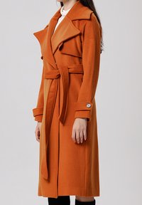 Manteau trench en mélange de laine orange avec une taille ceinturée, de larges revers et des poignets boutonnés, présentant une coupe longue et droite ainsi qu'une texture douce.