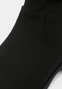 Bottine noire en matériau texturé semblable au daim, avec des accents cousus, une forme arrondie et une petite ouverture latérale.