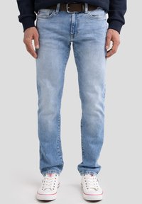 Mees, kes kannab helesiniseid teksapükse, pruuni vöö, valgeid tossusid ja tumedat pikakäist topi, seisab lihtsa taustaga ees.