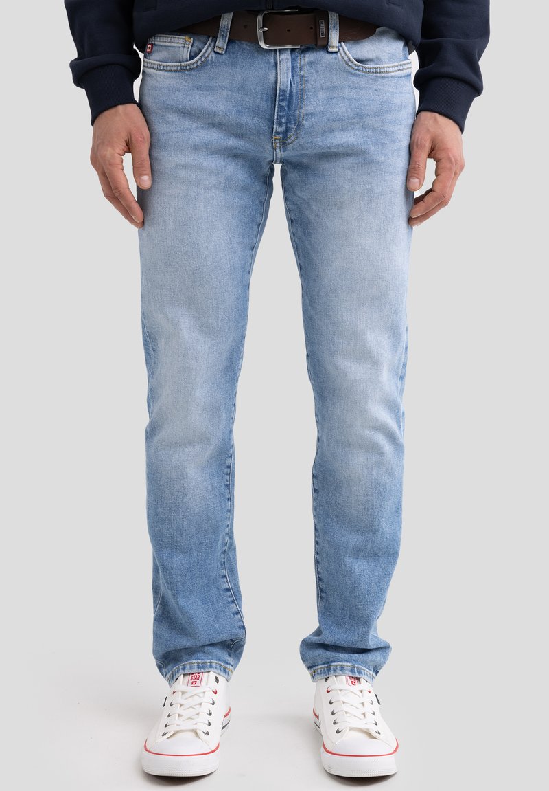 Mees, kes kannab helesiniseid teksapükse, pruuni vöö, valgeid tossusid ja tumedat pikakäist topi, seisab lihtsa taustaga ees.