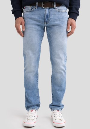 Férfi világoskék farmernadrágban, barna övvel, fehér tornacipőben és sötét hosszú ujjú felsőben, egyszínű háttér előtt áll.