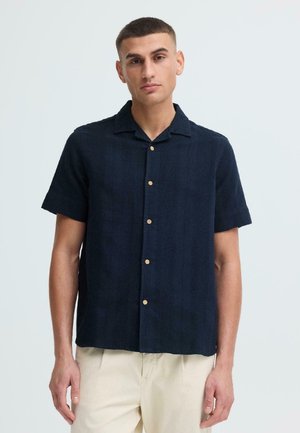 Man draagt een donkerblauw overhemd met korte mouwen en lichtbeige broek, staande tegen een effen lichte achtergrond.