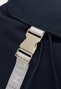 Detalle de una mochila de tela azul marino con un cierre en tono dorado y una correa gris claro que presenta texto en relieve, enfatizando la textura y el contraste de color.