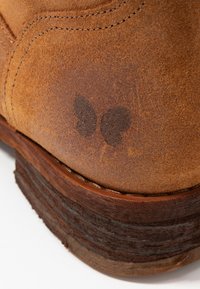 Primo piano del tacco di un stivale in suede marrone con una macchia d'acqua scura e cuciture visibili sulla superficie in pelle strutturata.