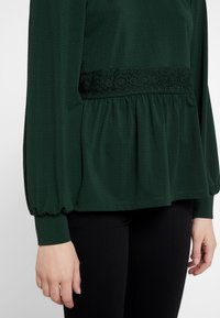 Top verde scuro con tessuto texturizzato, un accento in pizzo in vita e maniche lunghe e puffate. Abbinato a pantaloni neri, con un design peplum.