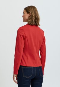 Röd figurnära jacka med långa ärmar, strukturerade axlar och hög halsringning. Tyget har en slät yta och kombineras med mörkblå jeans.