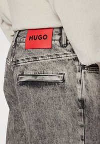 Flekkiintynyt musta denimfarkut, joissa on teksturoidun näköinen pinta. Takana punainen nahkainen merkintä, jossa lukee "HUGO", ja takataskun yksityiskohta.