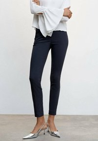 Pantalon noir ajusté de coupe slim, à la texture lisse et à la longueur chevilles, associé à des chaussures à talons argentées et un haut blanc à manches longues.