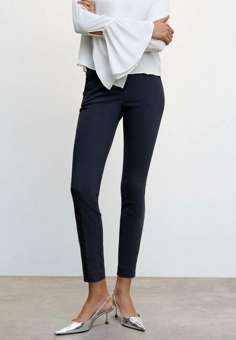 Pantalon noir ajusté de coupe slim, à la texture lisse et à la longueur chevilles, associé à des chaussures à talons argentées et un haut blanc à manches longues.