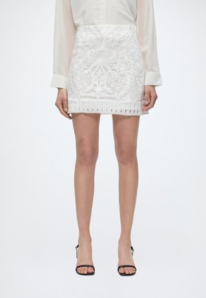 Λευκή κεντητή μίνι φούστα φορεμένη με κρεμ μπλούζα και μαύρα πέδιλα με ψηλό τακούνι και ανοιχτά δάχτυλα, εμφανιζόμενη από το μέσο του μηρού μέχρι τα πόδια.