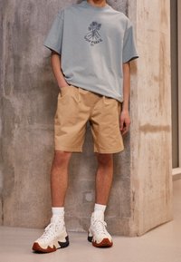 T-shirt azzurro chiaro con stampa grafica e pantaloni corti khaki. Sneakers alte bianche con suola in gomma nera e accenti marroni. Tessuti con texture lisce.