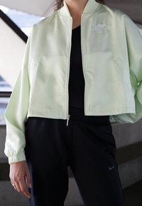 Ljusgrön kort jacka med dragkedja framtill, ribbade muddar och Nike-logotyp. Kombinerad med svarta byxor med en slät, mjuk yta.