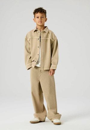 Junge steht und trägt eine beige übergroße Jacke und Hose mit weißen Turnschuhen vor einem schlichten hellen Hintergrund.