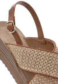 Tan braided wedge sandal featuring a woven upper, brown leather accents, adjustable strap, and textured sole.