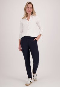Vrouw in een witte knopblouse, marineblauwe slim-fit broek en witte sneakers die loopt met één hand in de zak tegen een effen achtergrond.